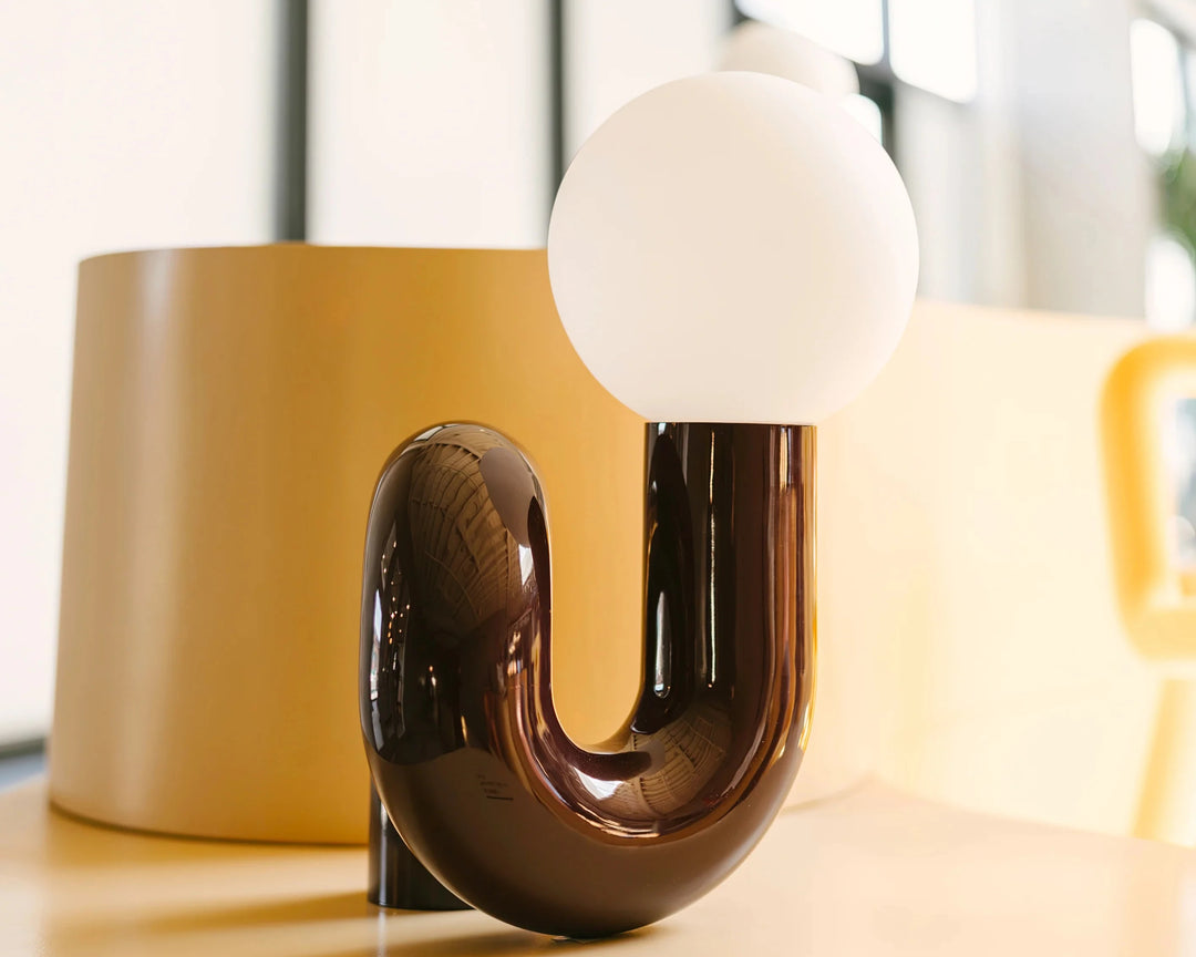 A close-up view of the Caterpillar Table Lamp featuring a glossy burgundy U-shaped base and a smooth white globe light, placed against a warm yellow-toned background in a modern indoor setting.