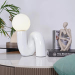 Load image into Gallery viewer, A Caterpillar Table Lamp with a glossy white curved base and softly glowing globe light displayed on a modern marble-topped table, accompanied by a neutral-toned abstract sculpture and stacked design books, creating a serene and stylish interior with greenery accents.
