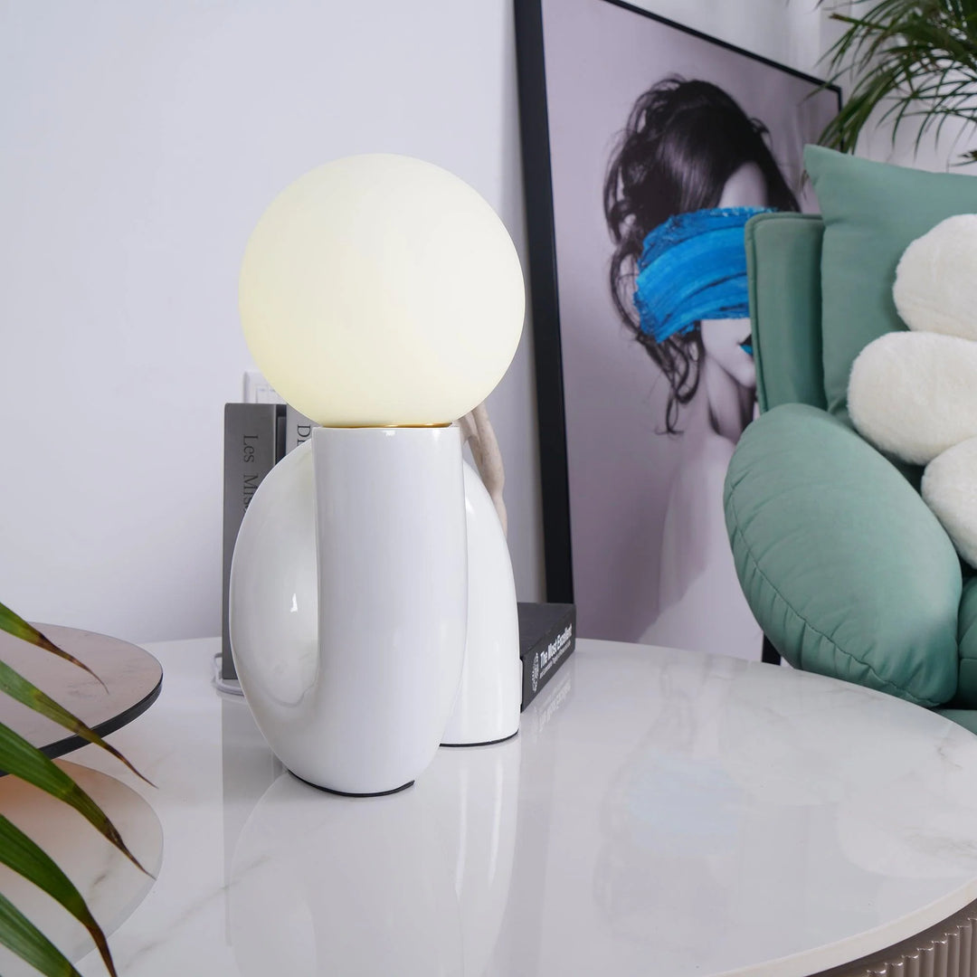 A Caterpillar Table Lamp with a glossy white curved base and glowing globe light placed on a sleek marble surface, styled with modern decor, a potted plant, and a framed abstract art print featuring a portrait with bold blue accents in a chic, contemporary living space.