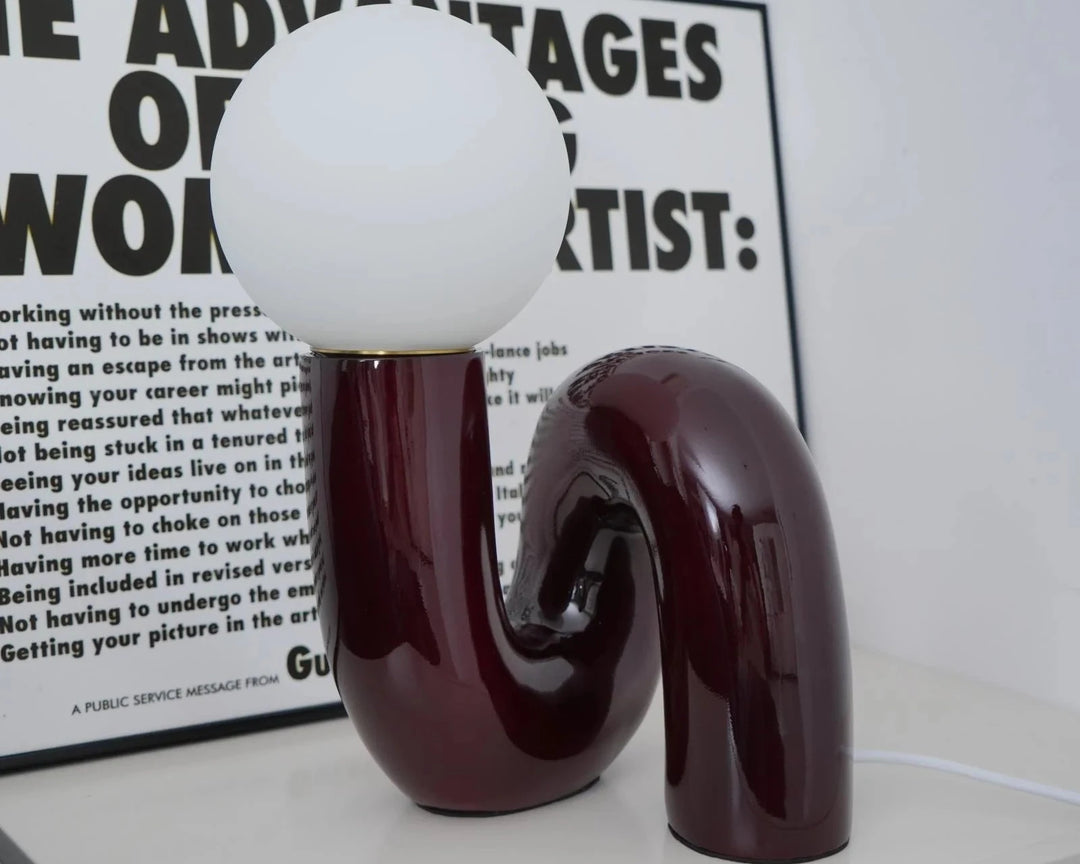 The Caterpillar Table Lamp with a glossy burgundy curved base and white globe light placed on a white surface, styled in front of a bold black and white typography art print in a modern minimalist room.