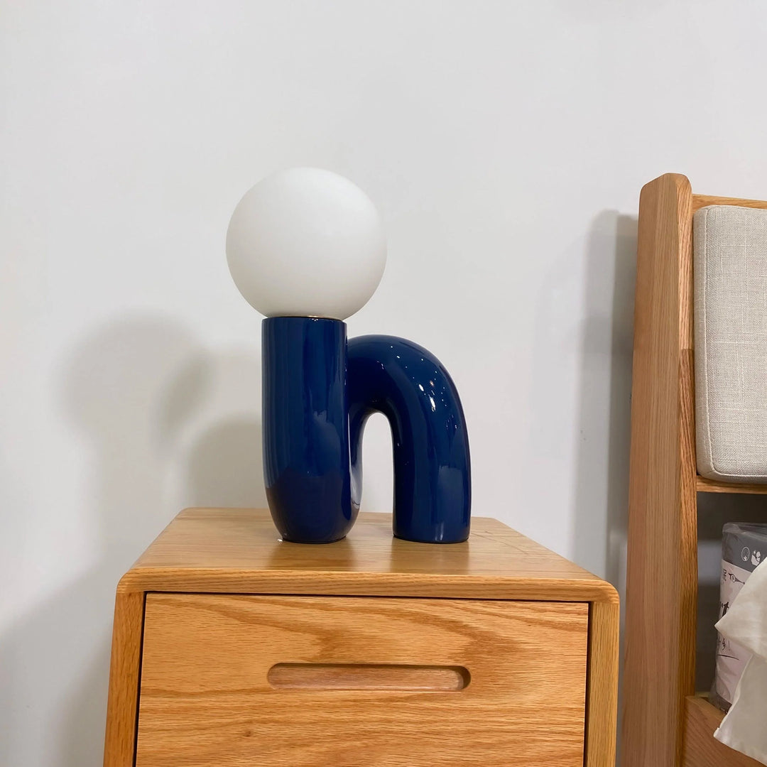 A Caterpillar Table Lamp with a glossy navy blue curved base and white globe light placed on a natural wood bedside table, styled in a cozy bedroom setting with a minimalist wooden shelf and neutral bedding.