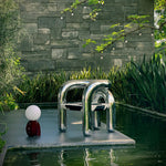 Load image into Gallery viewer, A modern Caterpillar Table Lamp with a burgundy curved base and white globe light placed beside a sleek metallic tubular chair on a platform surrounded by a tranquil pond, lush greenery, and hanging string lights in an elegant outdoor setting.
