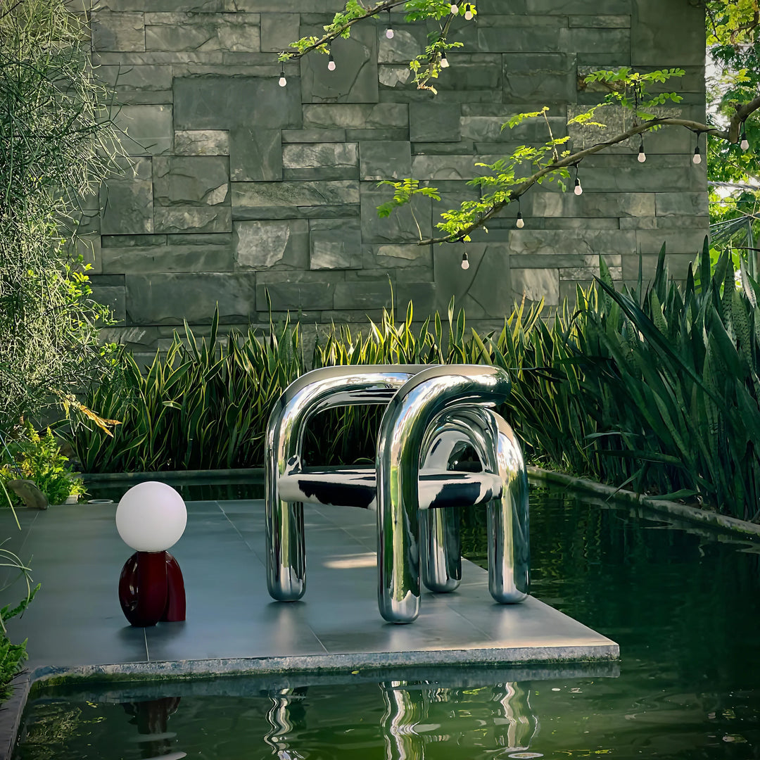 A modern Caterpillar Table Lamp with a burgundy curved base and white globe light placed beside a sleek metallic tubular chair on a platform surrounded by a tranquil pond, lush greenery, and hanging string lights in an elegant outdoor setting.