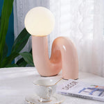 Load image into Gallery viewer, A Caterpillar Table Lamp with a glossy blush pink curved base and glowing white globe light displayed on a lace-covered table, styled with a teacup, a home decor magazine, and leafy green plants, creating a soft and elegant ambiance near a sunlit window with sheer curtains.
