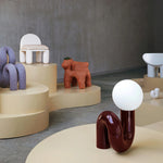 Load image into Gallery viewer, A modern and sculptural Caterpillar Table Lamp with a glossy burgundy curved base and a round white globe light, displayed in a contemporary showroom with artistic decor and furniture on soft beige podiums.
