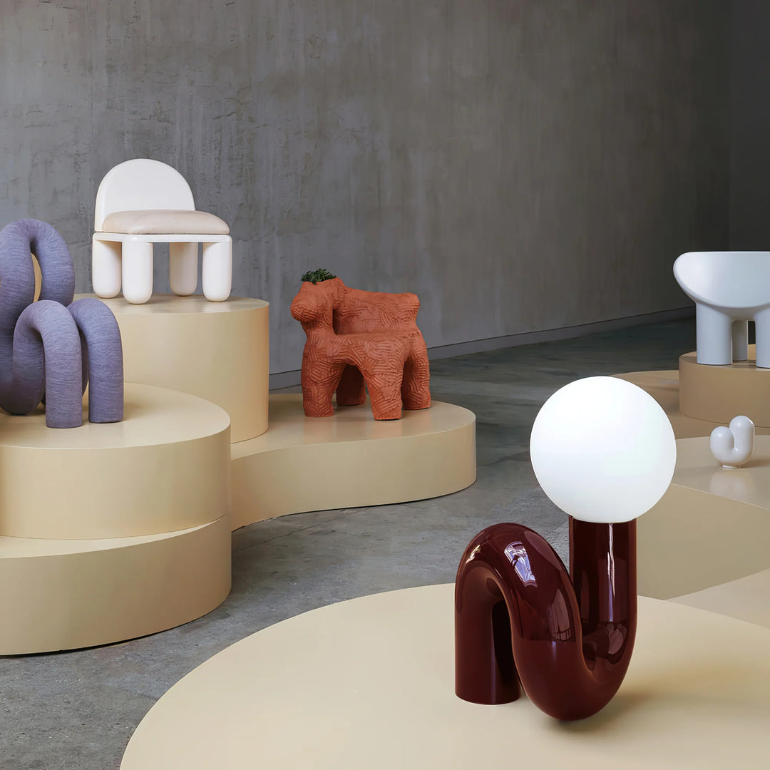 A modern and sculptural Caterpillar Table Lamp with a glossy burgundy curved base and a round white globe light, displayed in a contemporary showroom with artistic decor and furniture on soft beige podiums.