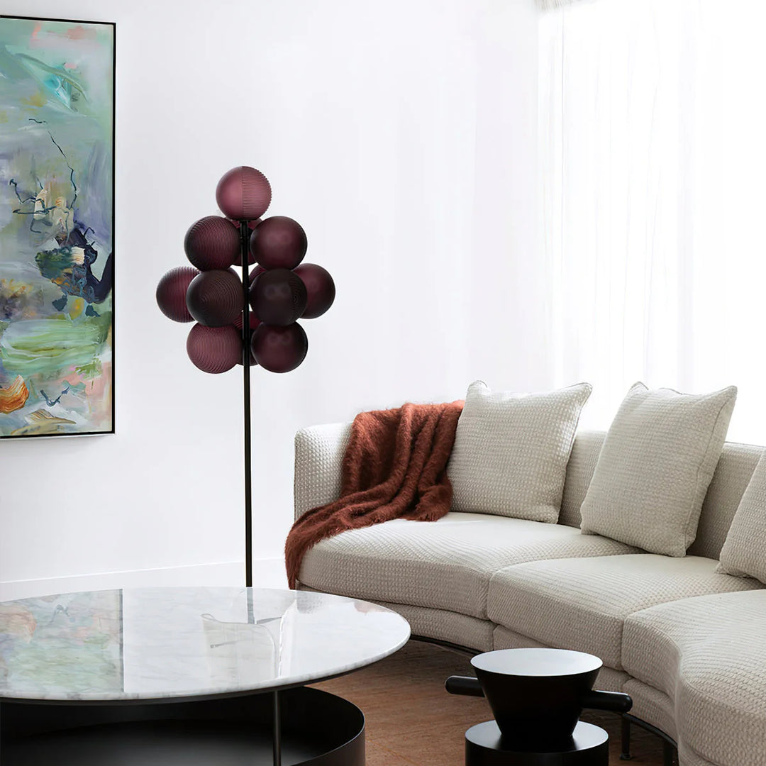 The Cépage Floor Lamp with burgundy globe-shaped lights on a black stand, placed in a bright living room featuring a white curved sectional sofa, a cozy rust-colored throw blanket, a round marble coffee table, and an abstract colorful wall painting for a modern artistic touch.