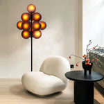 Load image into Gallery viewer, The Cépage Floor Lamp with glowing burgundy globes radiating warm amber light, placed beside a modern white boucle lounge chair and a black round side table styled with a minimalist vase holding branches with orange berries, creating a cozy and stylish reading nook.
