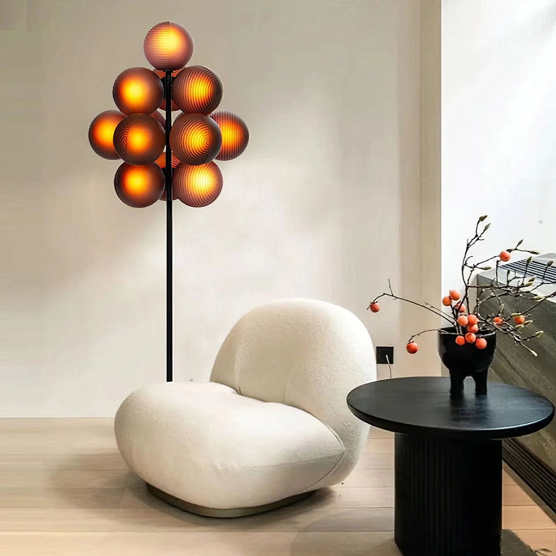 The Cépage Floor Lamp with glowing burgundy globes radiating warm amber light, placed beside a modern white boucle lounge chair and a black round side table styled with a minimalist vase holding branches with orange berries, creating a cozy and stylish reading nook.