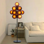 Load image into Gallery viewer, The Cépage Floor Lamp with glowing amber-toned burgundy globes on a sleek black stand, creating a warm ambiance in a minimalist living space featuring a neutral sectional sofa, a rattan side table with decor accents, framed art, and a potted plant for a serene and inviting atmosphere.
