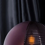 Load image into Gallery viewer, A close-up detail shot of the Cépage Floor Lamp showcasing the rich burgundy globe with ribbed glass texture, illuminated by a warm amber glow against a dramatic dark curtain background, highlighting its elegant and modern design.
