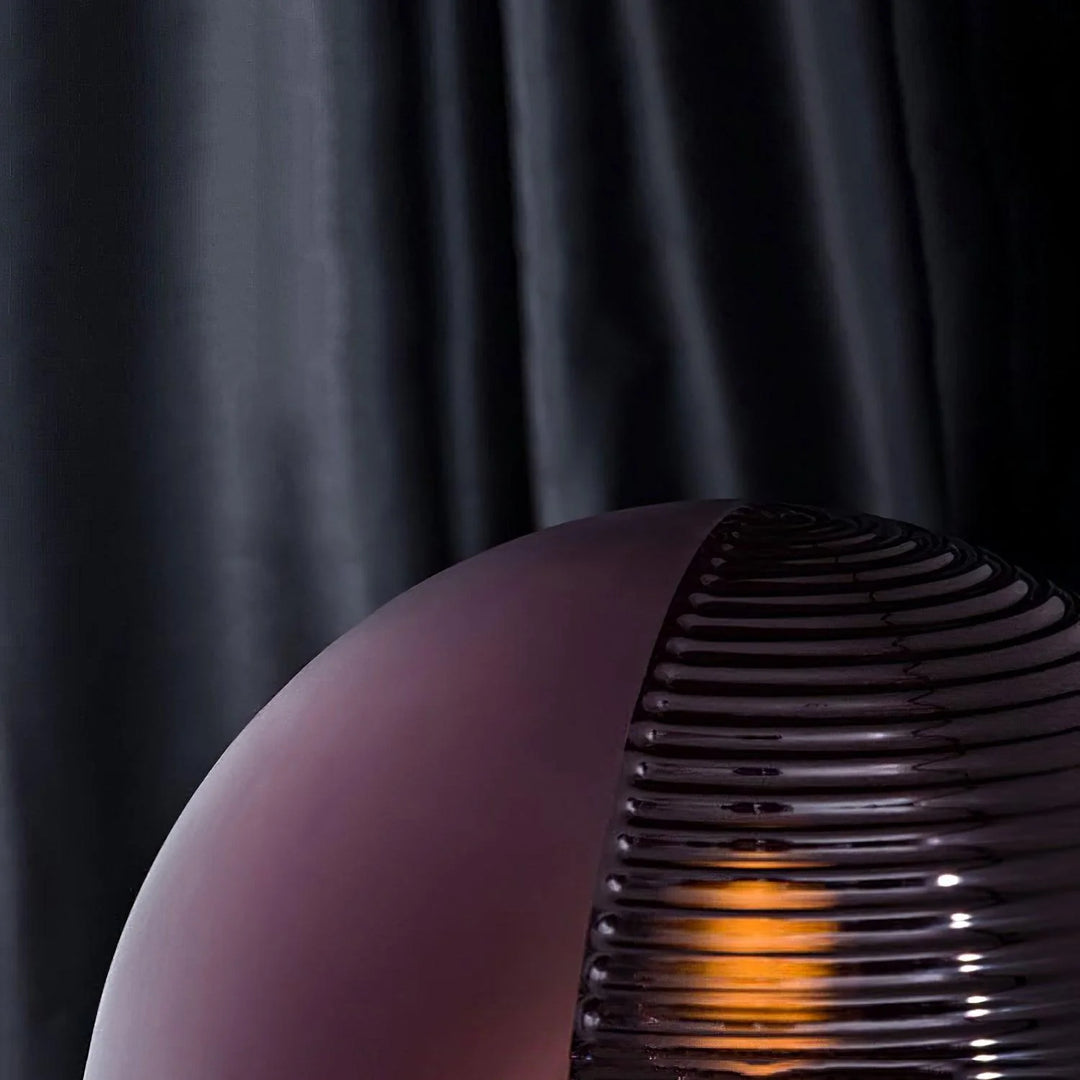 A close-up detail shot of the Cépage Floor Lamp showcasing the rich burgundy globe with ribbed glass texture, illuminated by a warm amber glow against a dramatic dark curtain background, highlighting its elegant and modern design.