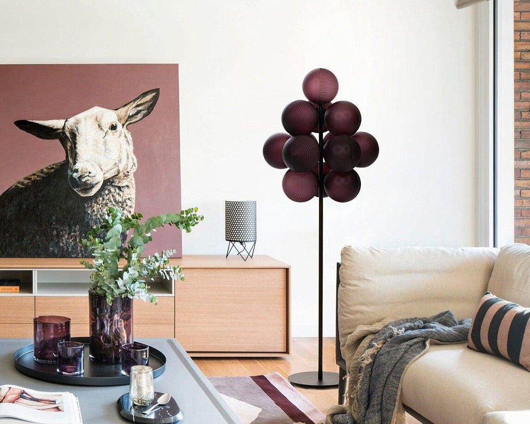 The Cépage Floor Lamp with burgundy globe-shaped lights on a black stand, styled in an eclectic living room featuring a neutral sofa with layered cushions, a modern coffee table with decorative glassware, a bold sheep art piece, and a light wood media console for a playful and sophisticated touch.