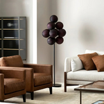 Load image into Gallery viewer, A contemporary Cépage Floor Lamp featuring a cluster of rich burgundy globe-shaped lights on a sleek black stand, elegantly placed in a modern living room with brown leather armchairs, a white sofa with brown accent pillows, and a glass-front cabinet.
