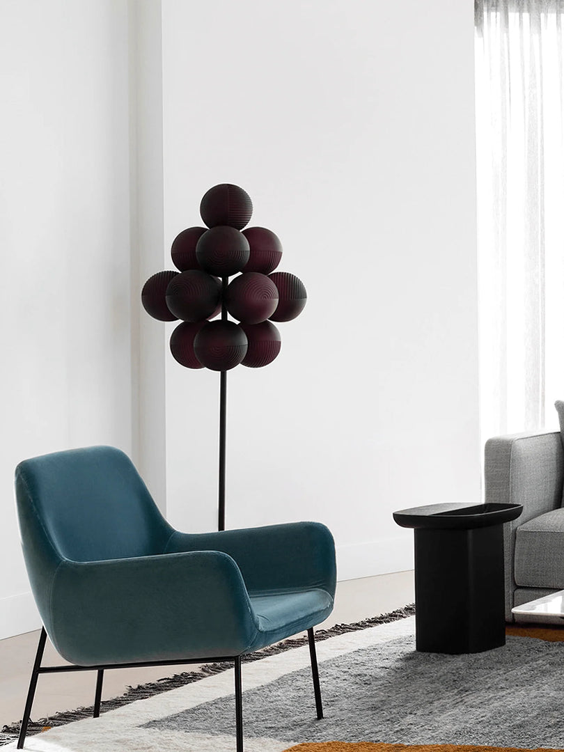 The Cépage Floor Lamp with deep burgundy globe-shaped lights on a black stand, styled in a modern lounge area featuring a teal mid-century armchair, a black side table, a gray sofa, and a textured area rug, creating a sleek and contemporary space.