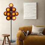 Load image into Gallery viewer, The Cépage Floor Lamp with glowing amber burgundy globes on a sleek black stand, adding a warm ambiance to a stylish living room featuring a mustard velvet sofa, a neutral textured throw pillow, a modern wooden stool, and an abstract white wall art piece for a cozy yet sophisticated look.
