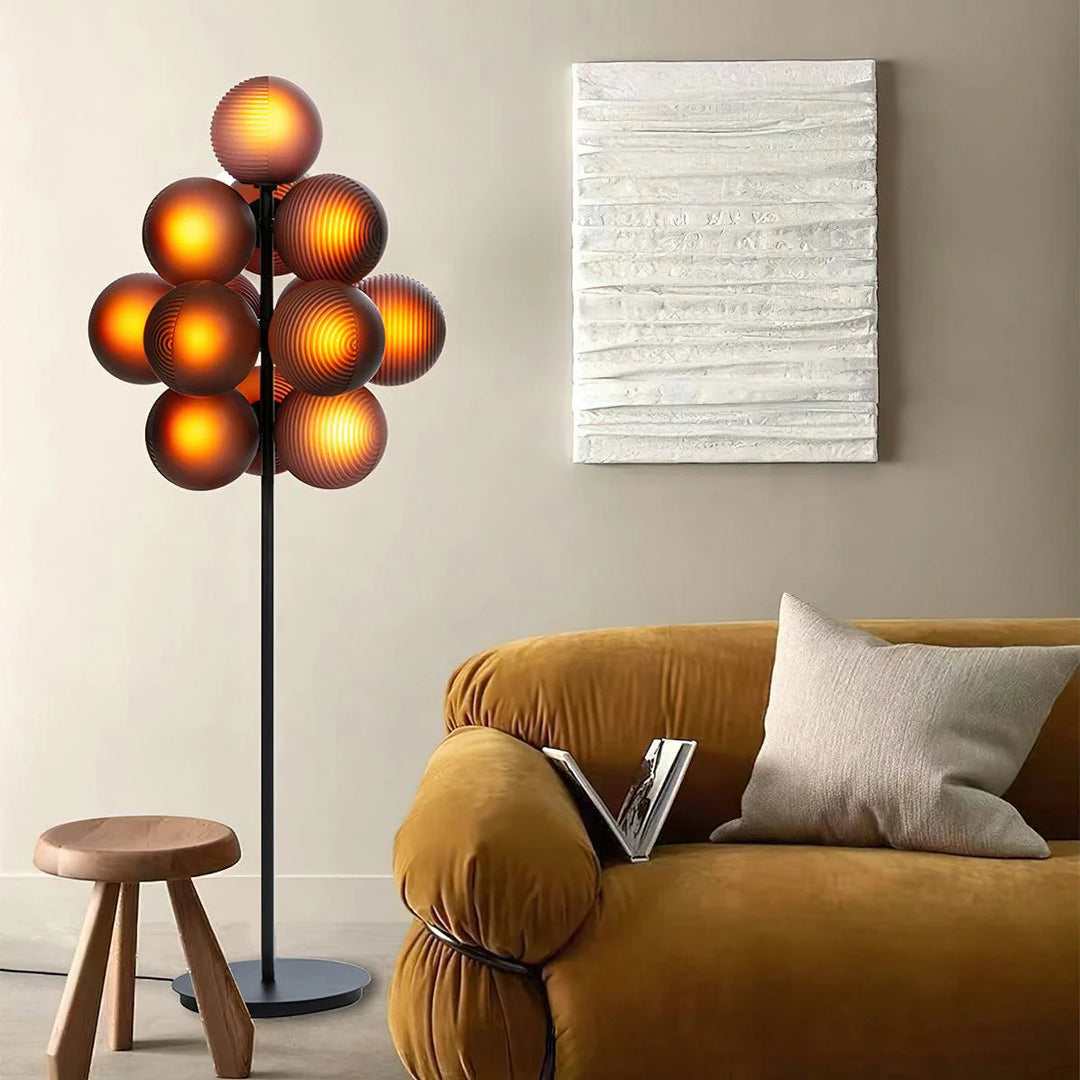 The Cépage Floor Lamp with glowing amber burgundy globes on a sleek black stand, adding a warm ambiance to a stylish living room featuring a mustard velvet sofa, a neutral textured throw pillow, a modern wooden stool, and an abstract white wall art piece for a cozy yet sophisticated look.