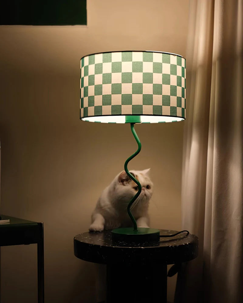 A cozy, softly lit nook featuring a Checkerboard Lamp with a green and white patterned shade and a whimsical spiraled green base. The lamp sits on a dark, speckled round side table, casting a warm glow, while a curious white cat rests beneath, adding a charming and playful touch to the serene ambiance of the room.