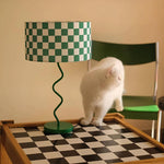 Load image into Gallery viewer, A delightful scene featuring the Checkerboard Theme Lamp with a green and white checkered shade and a twisted green base, playfully coordinated with a checkered table where a curious white cat is perched. The vibrant green chair in the background complements the lamp’s design, creating a lively and charming setting that adds character to any room.
