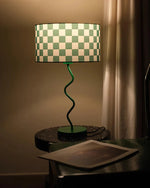 Load image into Gallery viewer, A chic and minimalist setting showcasing a Checkerboard Lamp with a green and white checkered shade and a spiral green base, emitting a warm glow. The lamp is placed on a sleek, speckled side table beside a draped curtain, creating a cozy ambiance. A nearby green chair holds an open magazine, adding a touch of casual elegance to the scene.
