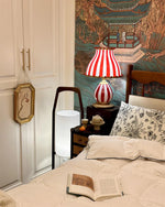 Load image into Gallery viewer, The Circus Table Lamp with red and white stripes and pom-pom accents, styled on a bedside table alongside teacups and a reading book, set against an ornate Asian-inspired wall mural and complemented by cozy bedding with floral patterns, creating a warm and eclectic bedroom ambiance.
