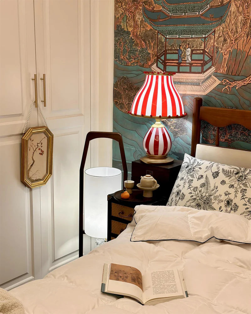 The Circus Table Lamp with red and white stripes and pom-pom accents, styled on a bedside table alongside teacups and a reading book, set against an ornate Asian-inspired wall mural and complemented by cozy bedding with floral patterns, creating a warm and eclectic bedroom ambiance.