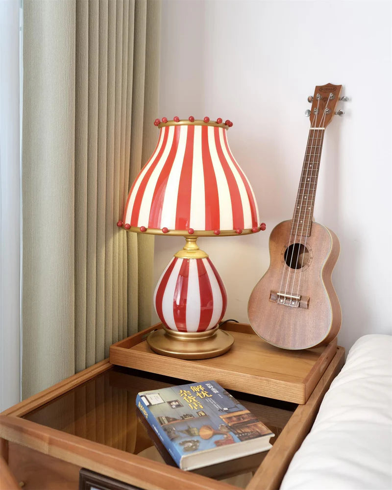 The Circus Table Lamp with a red and white striped shade and pom-pom accents, placed on a wooden bedside table with a ukulele and a book, creating a cozy and inviting bedroom setup with soft lighting and natural wood tones.