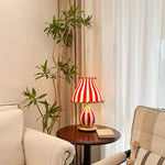 Load image into Gallery viewer, The Circus Table Lamp with red and white stripes and playful pom-pom details, placed on a dark wooden side table beside a cream armchair and a tall indoor plant, creating a cozy and inviting reading nook with soft natural light filtering through sheer curtains.
