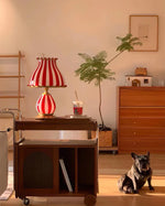 Load image into Gallery viewer, The Circus Table Lamp with a red and white striped design and pom-pom accents, placed on a wooden bar cart in a warm, sunlit living space, styled with greenery, a framed artwork, and a relaxed French bulldog lounging on the floor, creating a charming and inviting ambiance.
