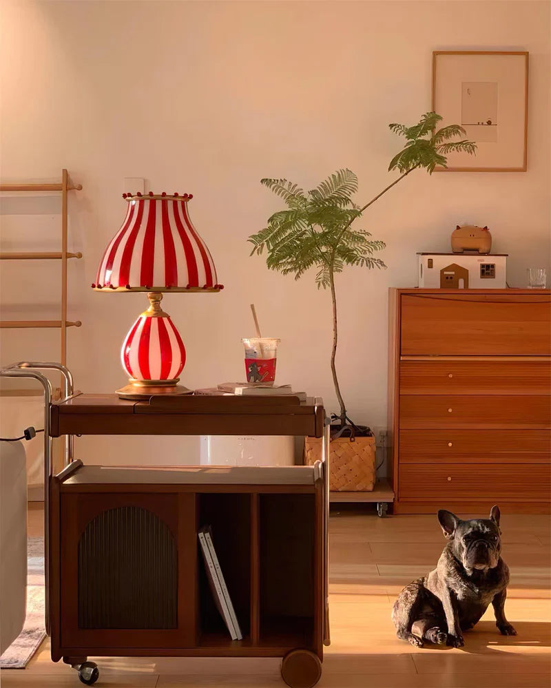 The Circus Table Lamp with a red and white striped design and pom-pom accents, placed on a wooden bar cart in a warm, sunlit living space, styled with greenery, a framed artwork, and a relaxed French bulldog lounging on the floor, creating a charming and inviting ambiance.