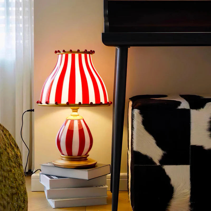 The Circus Table Lamp with a red and white striped shade and base, accented with pom-poms, styled in a cozy corner placed on stacked books next to a black table and cowhide-patterned ottoman, creating a playful yet chic interior setting with bold textures and patterns.