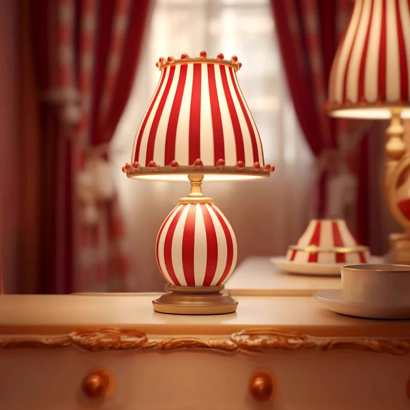 The Circus Table Lamp with a red and white striped shade and pom-pom accents, glowing warmly on an ornate cream vanity with gold detailing, styled with a teacup and surrounded by rich red drapery, creating a luxurious and whimsical atmosphere.