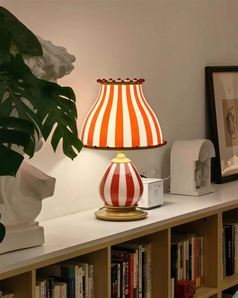 The Circus Table Lamp with a red and white striped shade and pom-pom accents, glowing warmly on a minimalist white shelf styled with art books, a framed print, and a decorative bust sculpture, creating a sophisticated yet whimsical home decor vignette.