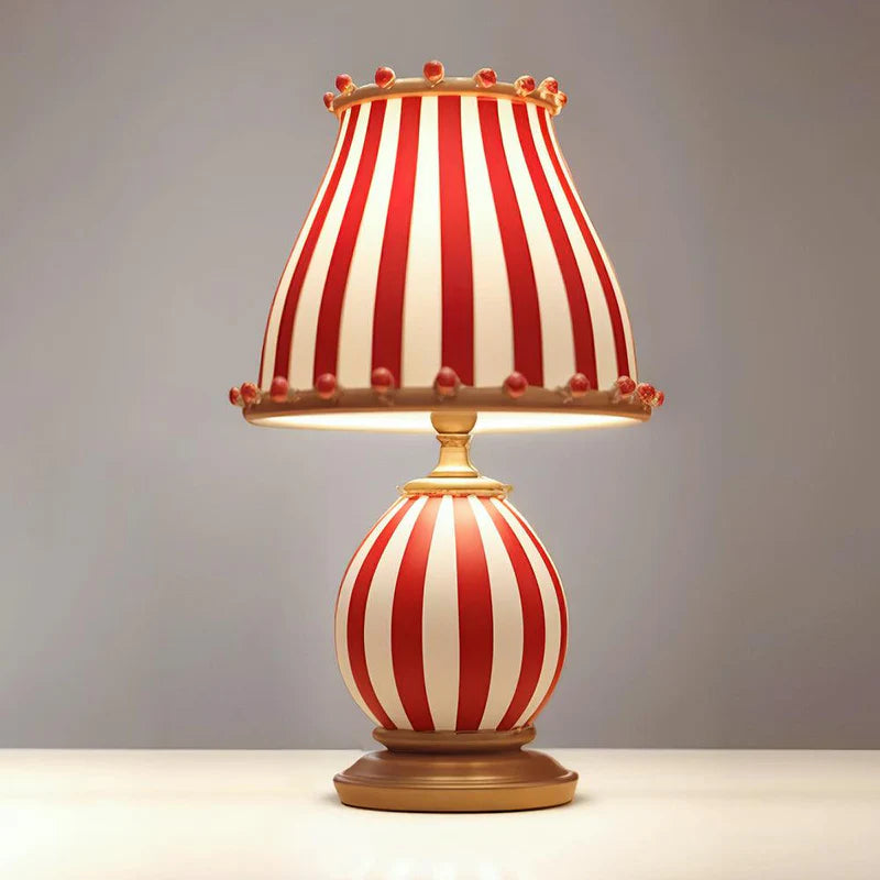 A whimsical Circus Table Lamp with a bold red and white striped design featuring a rounded base and matching lampshade, accented with playful pom-poms, placed against a neutral background to highlight its vintage-inspired charm and unique aesthetic.