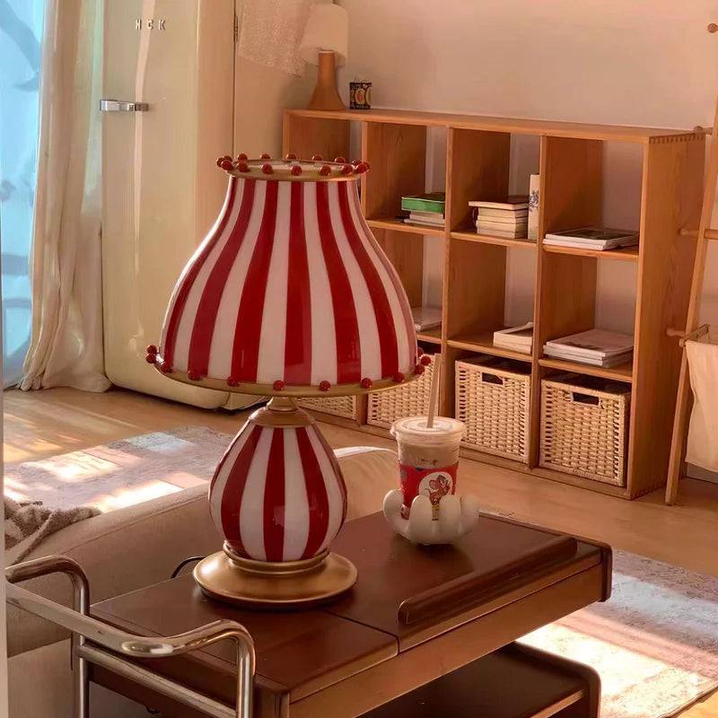 The Circus Table Lamp with a red and white striped shade and pom-pom accents, placed on a stylish bar cart in a sunlit living area with wooden shelving, woven baskets, and a cozy rug, creating a warm and inviting space filled with natural light.