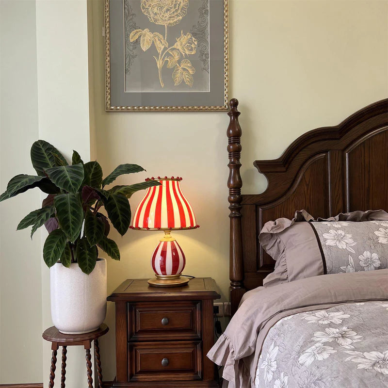 The Circus Table Lamp with a red and white striped shade and pom-pom accents, glowing warmly on a dark wood nightstand next to a classic wooden bedframe with floral bedding, complemented by a large leafy plant and framed botanical artwork, creating a charming vintage-inspired bedroom ambiance.