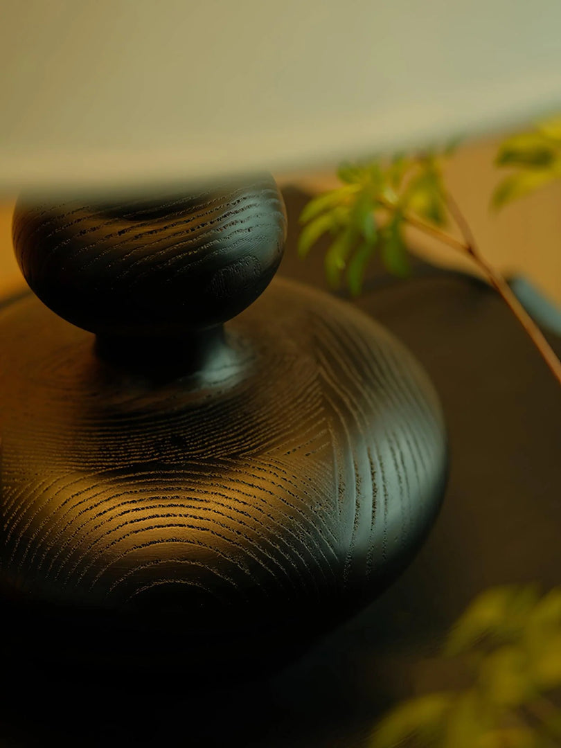 A close-up view capturing the intricate detail of a Wooden Gourd Lamp base, highlighting its unique textured surface and rich, dark finish. The soft ambient light accentuates the fine lines and patterns carved into the wood, paired with a subtle backdrop of soft green foliage, adding depth and a natural element to this artistic vignette.