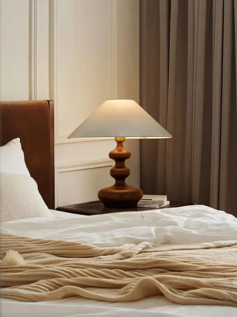 A serene bedroom setting illuminated by a Cozy Bedroom Gourd Lamp with a wooden base and classic lampshade, placed on a bedside table beside a plush bed with a knitted cream throw. The soft, warm lighting and elegant drapery contribute to a calm and inviting atmosphere.