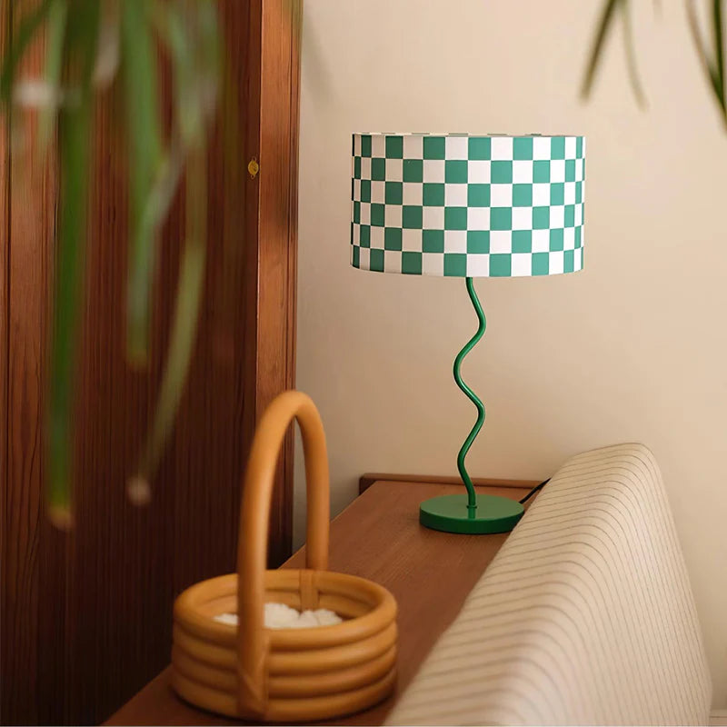 A cozy interior corner featuring a Checker Lamp with a green and white checkered shade and a whimsical twisted green base, set on a warm wooden surface. A stylish wicker basket adds a natural touch to the decor, while the surrounding wood accents and soft, neutral tones create a harmonious and inviting space. Lush greenery in the foreground enhances the organic and serene atmosphere.