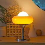 Load image into Gallery viewer, Crostata Table Lamp casting a warm ambient glow, featuring a glowing white orb within a vibrant orange glass bowl on a polished chrome base, styled on a wooden surface with decorative elements like dried flowers and a woven basket accent.
