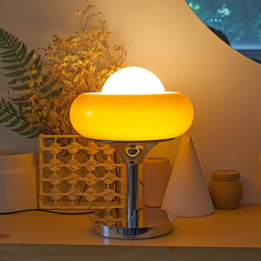 Crostata Table Lamp casting a warm ambient glow, featuring a glowing white orb within a vibrant orange glass bowl on a polished chrome base, styled on a wooden surface with decorative elements like dried flowers and a woven basket accent.