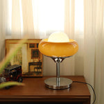 Load image into Gallery viewer, Crostata Table Lamp with a glowing white orb encased in an orange glass bowl, placed on a wooden surface alongside vintage decor and framed artwork, casting a warm glow that enhances the cozy and artistic ambiance.
