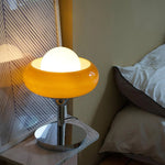 Load image into Gallery viewer, Crostata Table Lamp placed on a marble nightstand, featuring a glowing orb encased in an orange glass bowl, creating a cozy and ambient lighting effect in a modern bedroom setting.
