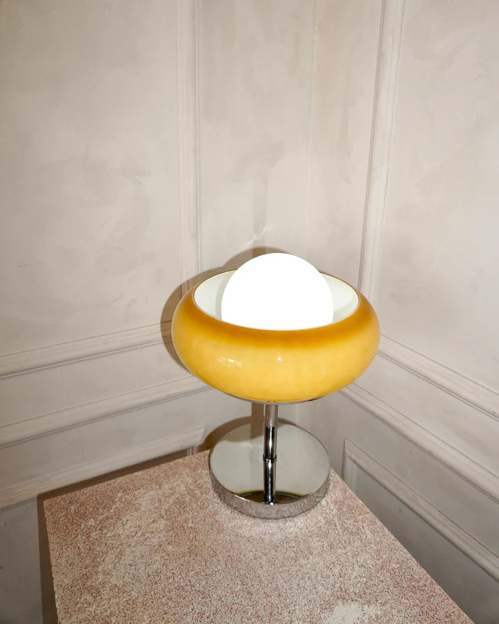 Close-up view of the Crostata Table Lamp displaying its polished chrome base, glowing white orb, and vibrant orange glass bowl, placed on a textured pedestal within an elegant neutral space.