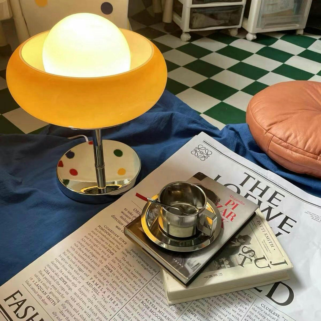 Crostata Table Lamp on a casual setup with a glowing white orb encased in a vibrant orange glass bowl, styled on a table with magazines, a coffee cup, and a cozy background, creating a warm and relaxed ambiance.