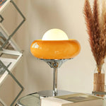 Load image into Gallery viewer, Crostata Table Lamp placed on a glass side table, featuring a glowing white orb inside a bold orange glass bowl with a polished chrome base, styled alongside decorative pampas grass and modern furnishings in a cozy interior setting.
