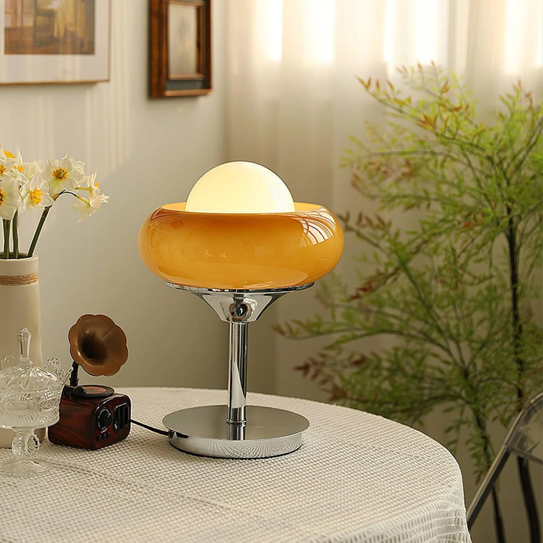 Crostata Table Lamp with a glowing white orb and vibrant orange glass bowl, styled on a round table with delicate decor, including fresh flowers and vintage accents, set against a backdrop of soft curtains and greenery.