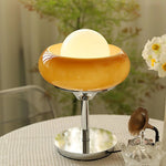 Load image into Gallery viewer, Crostata Table Lamp with a glowing white orb inside a glossy orange glass bowl and a polished chrome base, placed on a round table adorned with delicate decor and surrounded by greenery, creating a serene and elegant ambiance.
