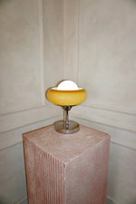 Load image into Gallery viewer, Crostata Table Lamp with a glowing white orb and orange glass bowl, elegantly showcased on a textured pedestal against a neutral-toned wall, creating a sophisticated and gallery-like presentation.
