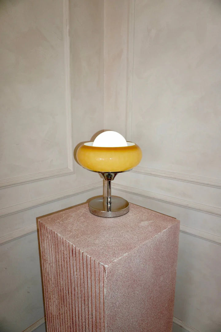 Crostata Table Lamp with a glowing white orb and orange glass bowl, elegantly showcased on a textured pedestal against a neutral-toned wall, creating a sophisticated and gallery-like presentation.